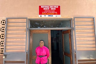 Esnart is wearing a bright pink blouse, and is standing outside of the maternal and child health ward, which is a beige pink building with a bright red sign.