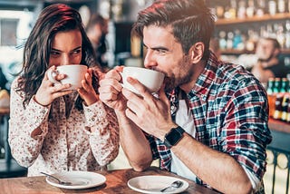 Two adults sitting in a coffee shop, drinking coffee. They are sat very close together, and smiling at each other flirtatiously over their coffee cups as they drink.