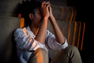 A Black woman leaning against a couch with her hands up to her face. She is wearing a casual blue shirt with brown slacks.