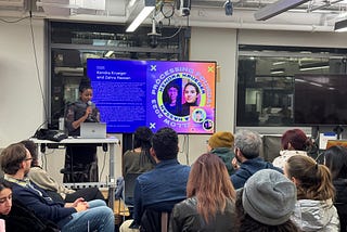 An image of our Programs Manager, Tsige Tafesse, facilitating and hosting the Fellow presentation at NYU ITP. She a dark skinned Ethiopian woman with braided hair tied up into a bun. The T.V. screen shows an image of Kendra Krueger and Zahra Hassan’s bio