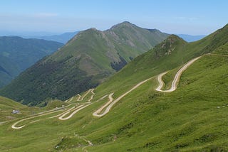 Green mountains under a blue sky, with a pale stone road zig-zagging up the mountain.
