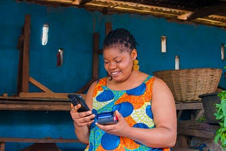 An African lady smiling while holding a phone in the right hand and a Point-of-Sale device in the left hand.