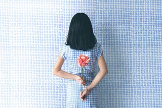 Back of a female-identified person holding a flower behind her back. She has shoulder length black hair. She is wearing a black and white dress, in front of a background that matches.