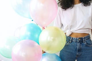 Woman wearing white top and jeans holding a bunch of pastel balloons