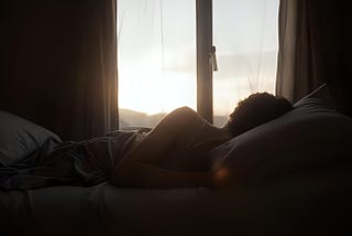 A woman laying in bed, on her side, in a dark room looking toward a window where she can see the tree tops in a distance and the sky to represent the feeling of being depressed.