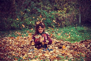 Young, brown haired woman with pony tail in a red sweater and fall colored scarf, sitting in a huge pile of Fall leaves, falling all around her in Sukhasana with prayer hands.