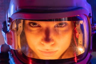 A woman wearing a hazmat helmet stares intensely into the camera, purple hues of light emanate off her helmet, and her face is framed in yellow light.
