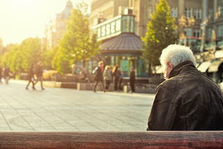 An elderly man with his back to the camera sits on a bench in a city square