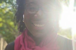 Selfie of the author, Scarlet Ibis James. She is backlit by brilliant sunshine and is wearing a red scarf. As usual, she has a dazzling smile.