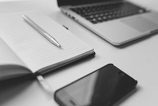 A laptop, phone, notebook, and pen laying on a desk, all in monochrome