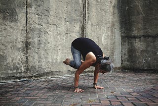 Mid aged woman doing the crow pose on a brick floor surrounded by concrete walls.