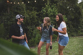 Two white women in cut off jeans and t-shirts talk to a white man in jeans and open shirt with a t-shirt underneath, also holding a drink. They are outside in a back garden, standing on grass. A string of out door lights hangs behind them. In the foreground the man’s lower torso is obscured by a bush, giving the impression that we are looking into the garden.