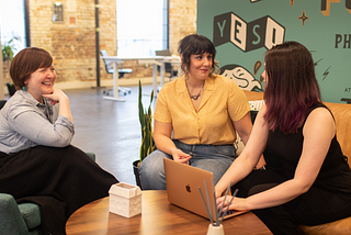 Jen, Sara, and Emily sit at a table in front of a teal mural wall. Jen has very short brown hair, a blue shirt and black skirt. Sara is wearing jeans and a yellow shirt. Emily is working on a laptop and wearing a black jumpsuit.