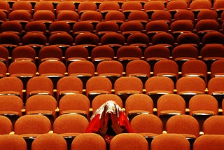 A single audience member sits in a big empty theater of orange seats. The audience member is hiding their head under their coat.