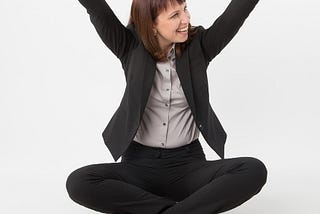 Woman in work attire — suit, and blouse — sits cross legged in front of a laptop and puts both arms in the air to celebrate