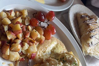 A vertical photo of many food items from the Uptown establishment Crepes and Grapes. This includes an omelet, potatoes, a crepe, and grapes!