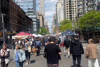 People walking on a pedestrianised avenue in New York City. It’s a beautiful day and the road is closed to cards because of a street fair. You can see the stalls on the street and many high rise buildings on either side of the road.