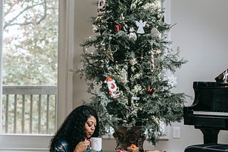 Black woman in pajamas enjoying drinking from a mug as she reads on the floor in front of her Christmas tree.