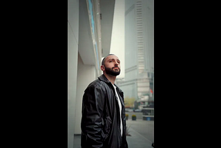A man with a closely shaved head looks upward while standing on a city street, tall glass buildings rising behind him.