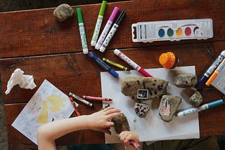 An aerial view of a markers and paints spread out on a wooden table. You can see a child’s hands painting on rocks and paper.