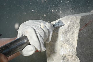 A white gloved left hand is holding a chisel to a piece of limestone. At the point where they meet, there is a cloud of stone chips and dust, as though the chisel has just been hit by a hammer.