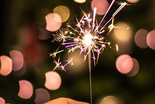 A hand holding out a single lit sparkler. Author used this image to connect to her celebration of one year on Medium.