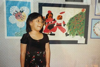 A photo of the author in as a young child, with a painting she created with herself and Santa (both with orange skin).
