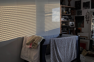 The image shows a baby travel bed in a dark room with light reflections from the window blinds on a white closet.