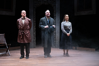 Three actors stand onstage and prepare to take a bow; the actor on the left wears a patterned smoking jacket, the one in the middle a gray suit, and the one on the right a blue skirt and floral blouse.