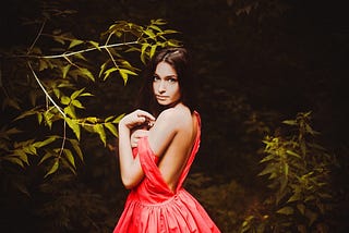 A beautiful young woman in a wooded area at night. She is standing with her back to the camera but has half-turned to face it. She has dark hair and pale skin. She is wearing a red, sleeveless ball gown, which is open to her waist at the back. She has slipped one shoulder of her gown halfway down her arm.