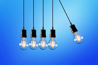 A blue background with 5 exposed Edison-type light bulbs hanging down on cords. All are light and the rightmost one is swung out as if it will knock in to the others, causing a chain reaction.