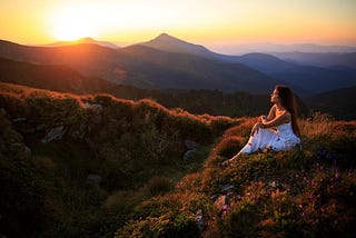 A lady in white dress sits on the earth staring at the setting sun