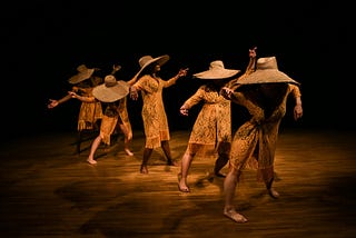 Five dancers standing in a diagonal row on a dim stage. They are wearing large woven hats and matching crochet dresses with arms out to their sides and looking in different directions.