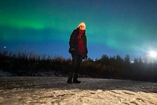 A person in a white hat and a black coat with a red scarf standing on the ground with snow. Behind are the Northern Lights and a bright full moon.