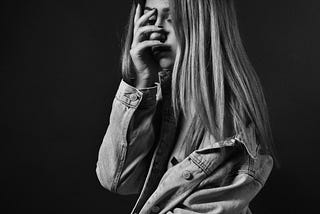 Woman in a denim jacket covering her face with her palm standing against an all-black background