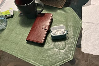 A green textured placemat is set on a table, displaying a few everyday items. On the left, there’s a dark-colored mug with what appears to be tea or coffee. Next to it is an opened packet of green tea, and a brown leather wallet-style phone case. On the right, a pair of AirPods Pro 2 earbuds rest inside an open, teal-colored charging case.