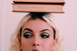 Woman with books on her head with blonde hair, dark roots, and bold eye makeup looking to the left.