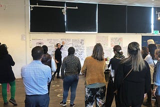 A group of people standing in a hall looking at a person speaking, pointing at a huge piece of paper covered in words and pictures