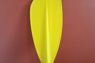 A bright yellow boat paddle leaning against a red wall