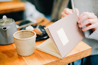 Table with gray tea pot, ceramic mug, and woman’s hands holding pen and an open brown journal.