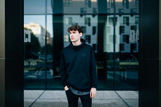 young man wearing dark clothing with hand in pocket standing in front of mirror glass building