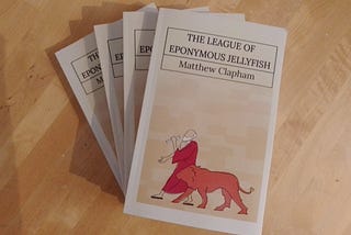 A number of paperback copies of the satirical SFF novel The League of Eponymous Jellyfish, by Matthew Clapham, sit on a desk, awaiting distribution to an eager global readership!