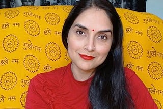 Indian woman with light brown skin, red bindi on her forehead, long black hair, red blouse, and saffron background with Sanskrit design printed on it.