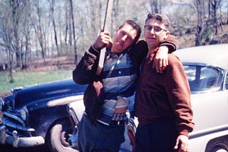 Two young men standing in front of their old cars, looking like they’re goofing around