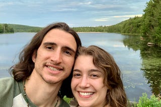 My partner and I smiling in front of a lake surrounded by green, bushy trees.