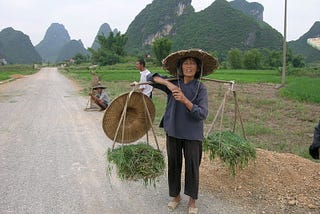 Three farmers on the road in Southern China’s Guangxi province. One is balancing two baskets filled with vegetables on pole. Fang-shaped karst-limestone hills in the background are found primarily in Southern China and Northern Vietnam.