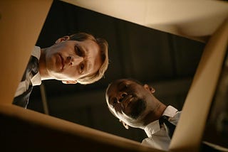 Two men in business attire are looking down into a cardboard box with serious and curious expressions. The image is taken from inside the box, showing their faces from a low angle.