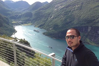 A picture of the author with a view of the blue water of the fjord and green mountains in the background