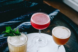 Three cocktails sitting on a white, marble circle. Cocktail on the left is gold in colour with a sprig of mint coming out the top, middle is a deep red cocktail in a coupe glass, and the right is a syrup-coloured cocktail in a nick and nora glass