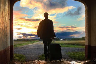 Photo of a man with a suitcase, silhoutetted in a large archway as he walks away, into a sunset. The walls in the foreground of the photo are dark, minimally lit, and he walks towards a blue and pink sun behind white nad grey clouds, a grass-lined road in his path.